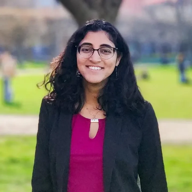 Headshot of Neha Presad on an outdoor background with a tree and a fence behind her.