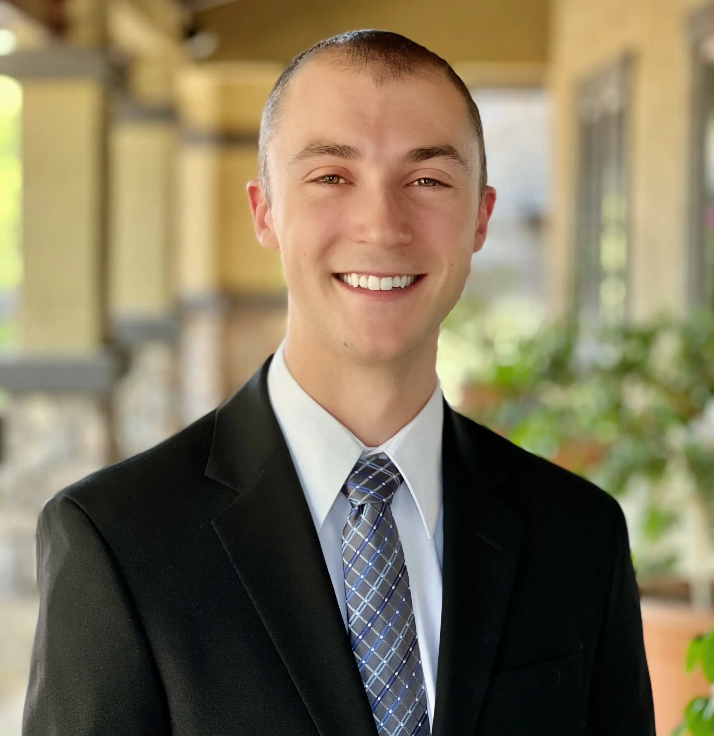 Headshot of Samuel Yates on an outdoor background with a terrace and a plant behind him.